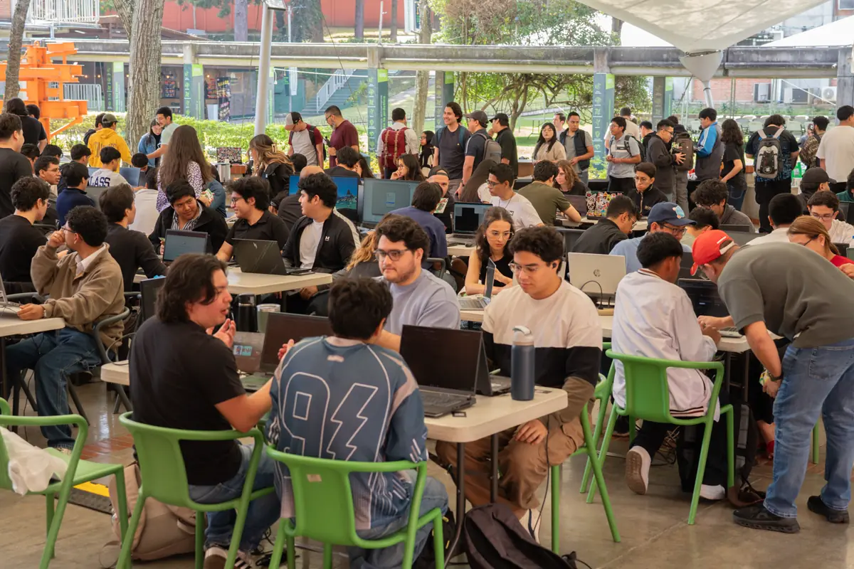 Personas con laptops en mesas bajo una carpa blanca, rodeadas de sillas verdes.