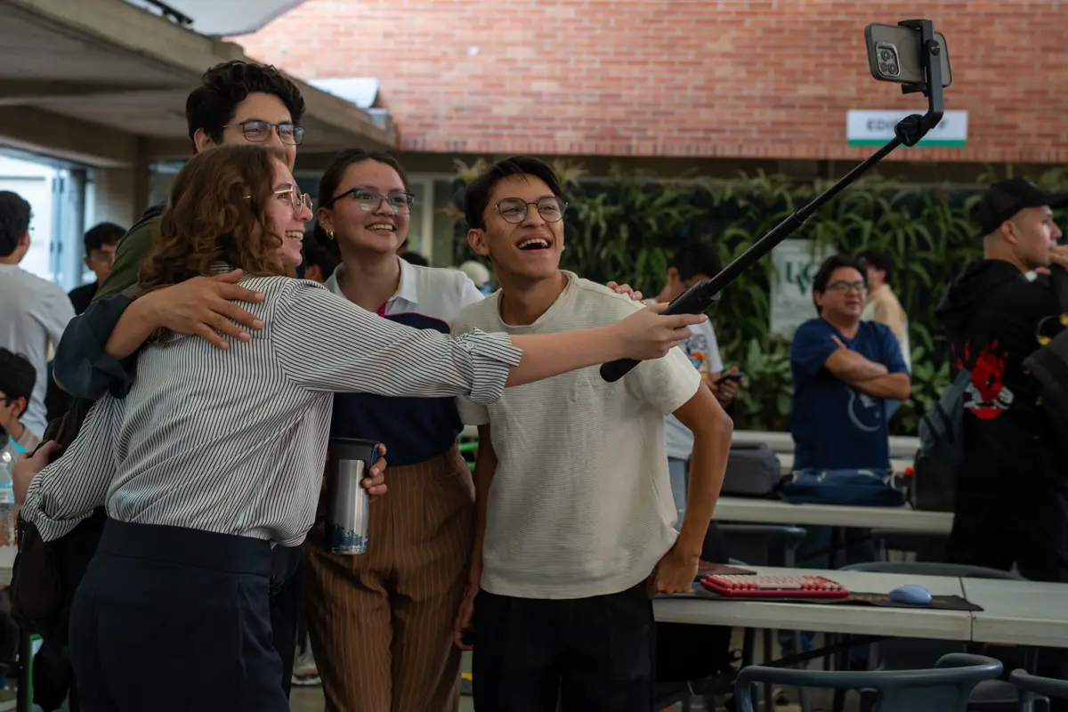 Cuatro personas sonríen mientras una sostiene un palo de selfie junto a una mesa.