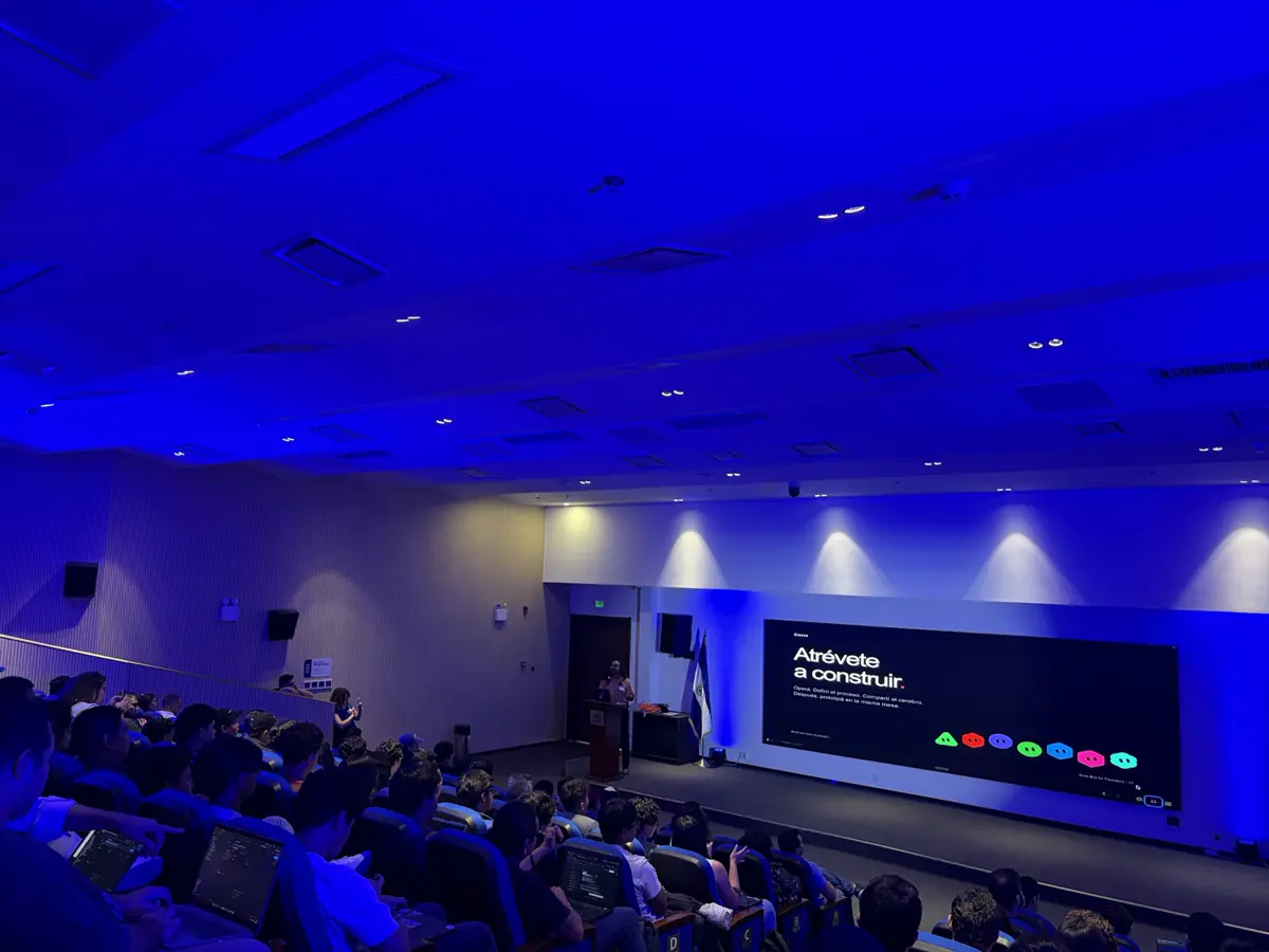 Auditorio en luz azul: una persona en el podio y la pantalla dice “Atrévete a construir”.