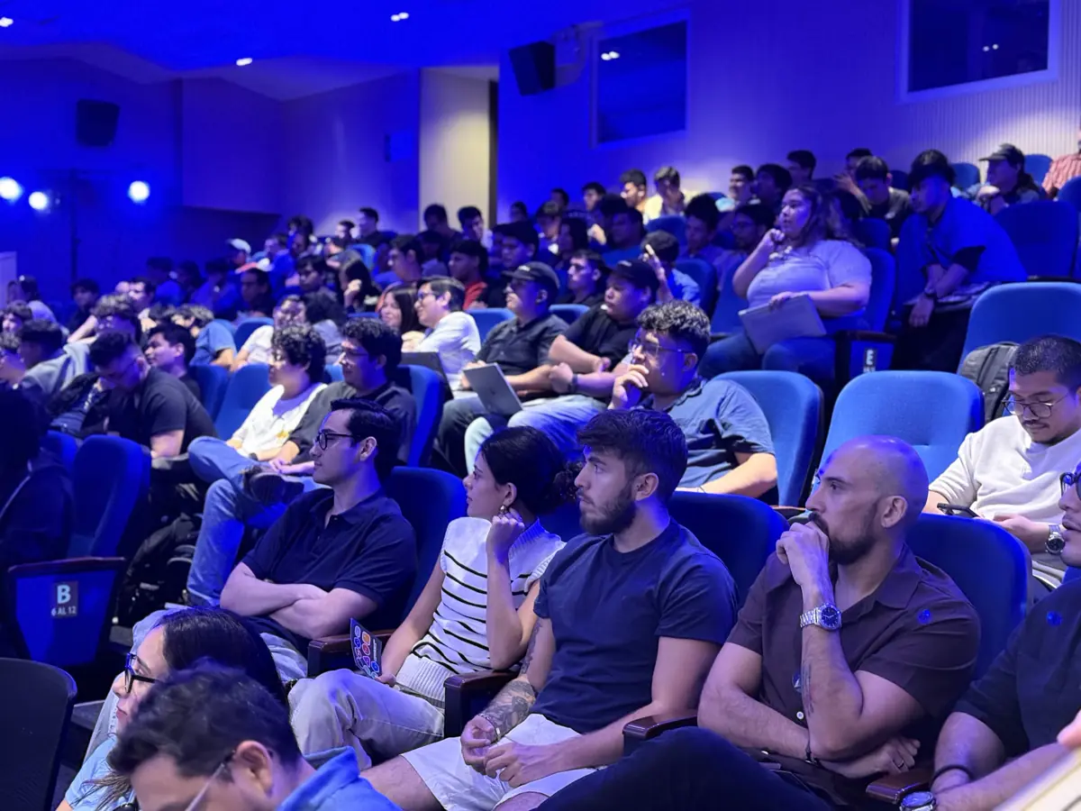 Filas de personas sentadas en butacas azules, bajo luz azul, mirando hacia el escenario.