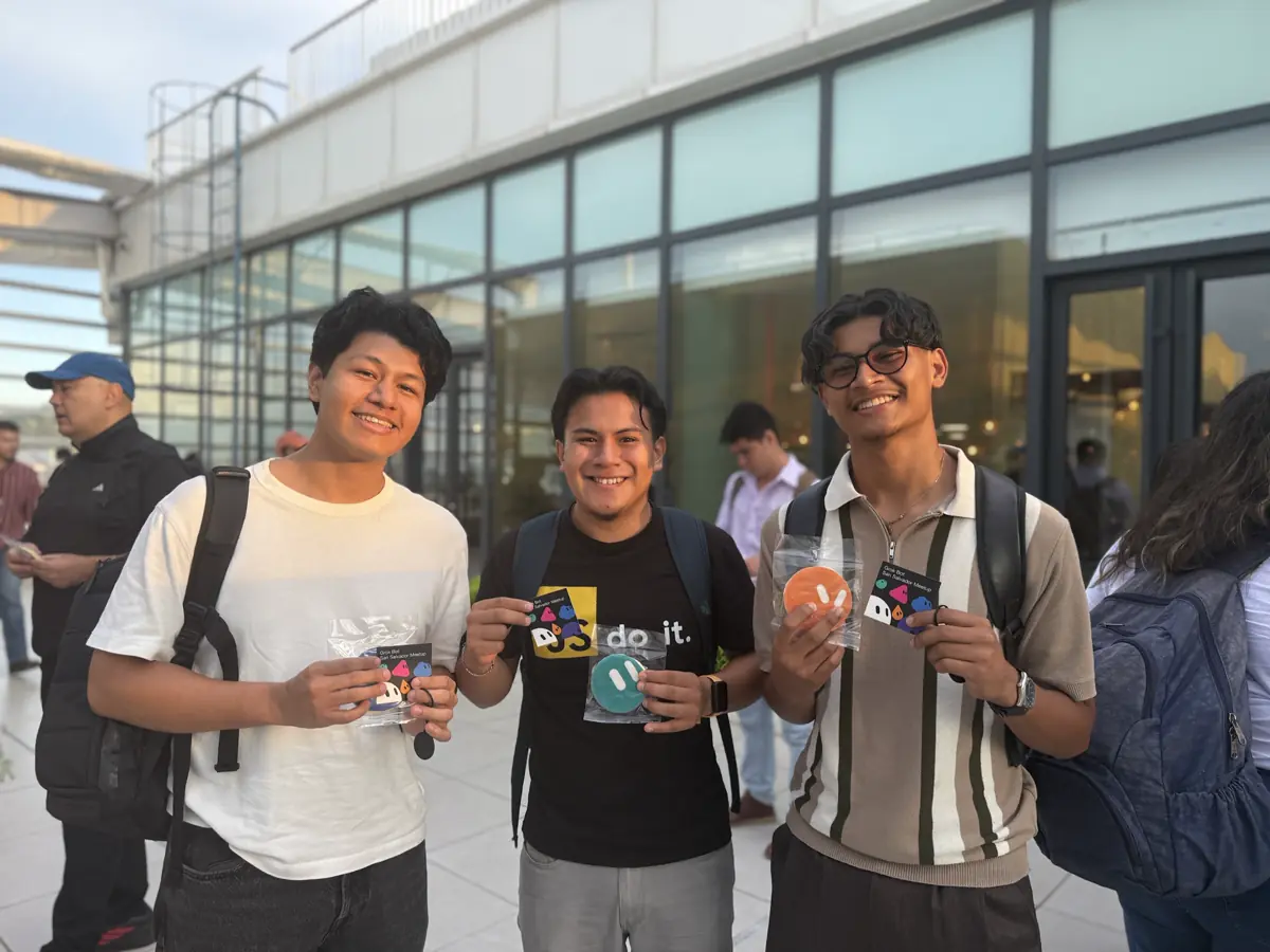 Tres personas sonríen afuera de un edificio de vidrio, mostrando stickers y tarjetas.