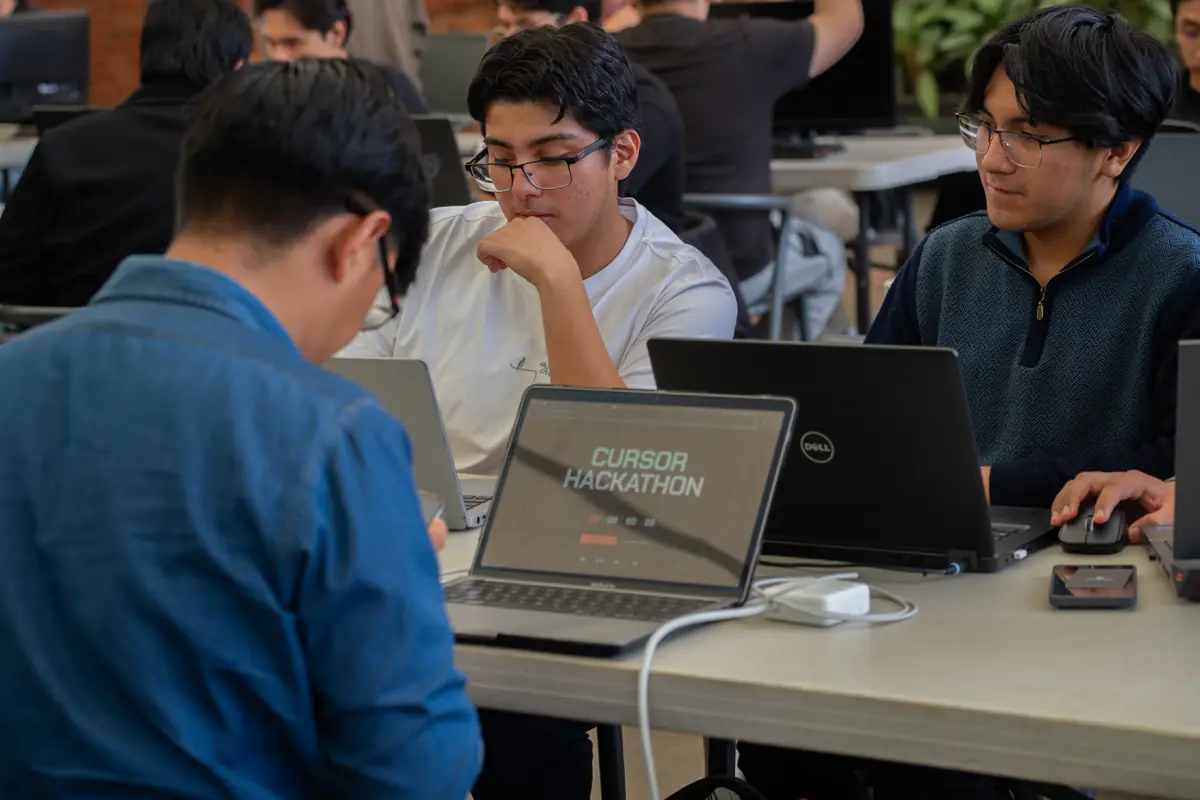 Tres personas alrededor de una mesa; la laptop del centro dice “Cursor Hackathon”.