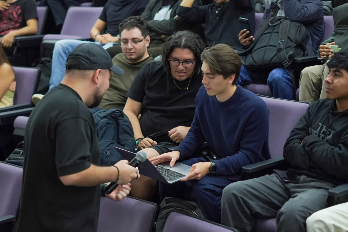 En butacas moradas, una persona con micrófono señala la laptop que sostiene otra.