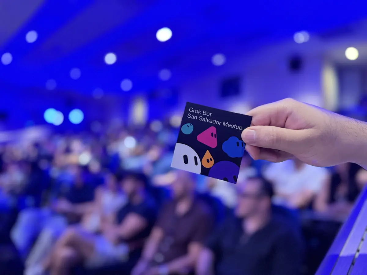 Una mano sostiene una tarjeta que dice “Grok Bot San Salvador Meetup”, con el público detrás.
