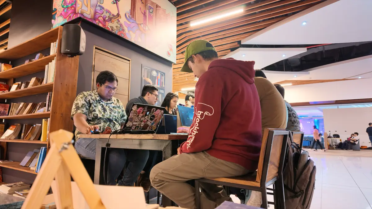 Personas con laptops alrededor de una mesa, junto a estantes de libros y un mural.