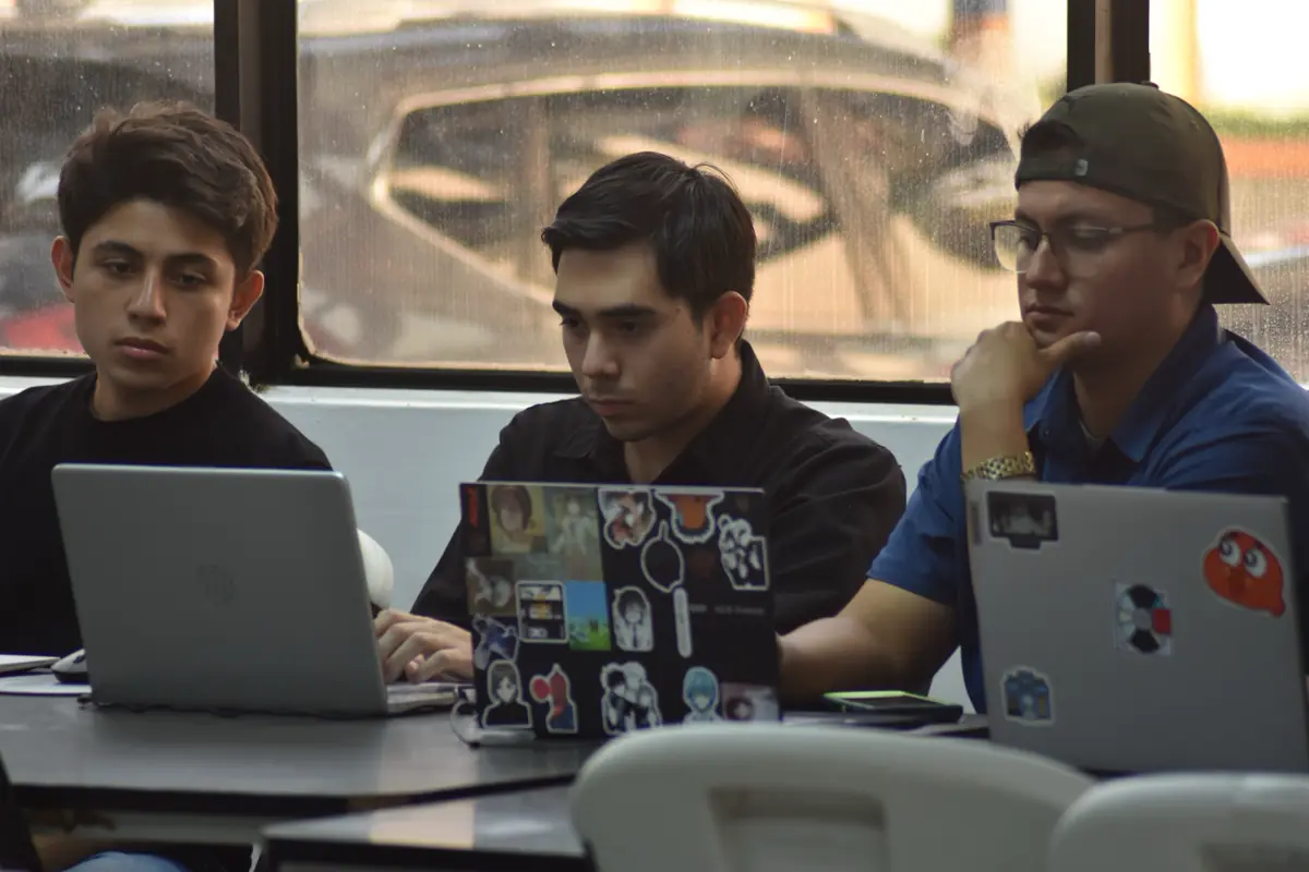 Tres personas trabajan en laptops junto a una ventana con el vidrio mojado.