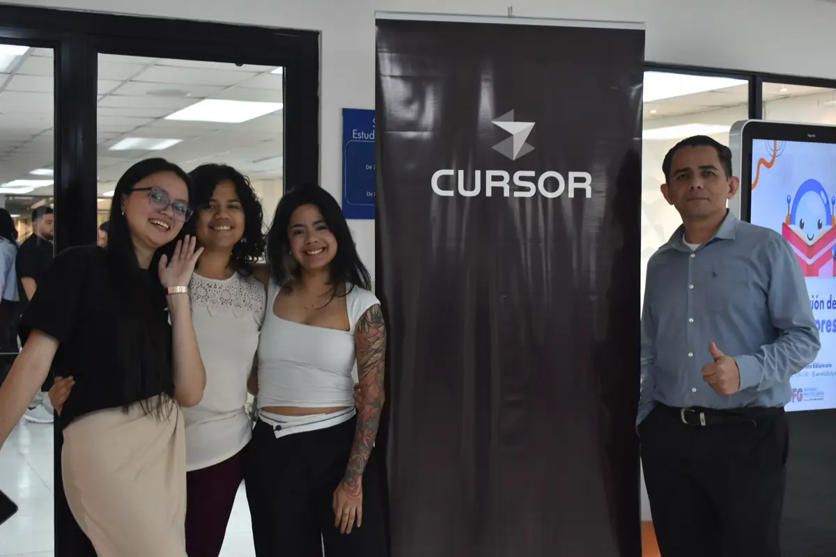 Cuatro personas posan junto a un banner vertical de Cursor, en un pasillo con vidrio.
